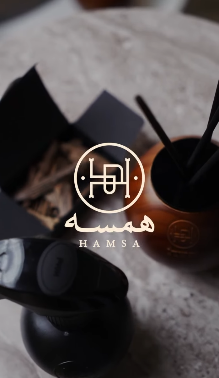Overhead flat-lay of a wooden diffuser sphere with reed sticks, matte black home spray bottles, and a dark gift box, with the Hamsa circular logo and Arabic wordmark overlaid at center
