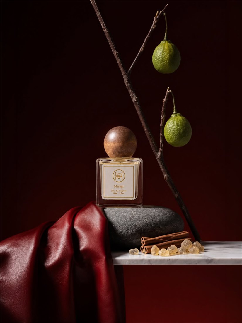 Mirage eau de parfum bottle with a wooden ball cap, styled with dried citrus, cinnamon sticks, and amber resin against a dark red backdrop