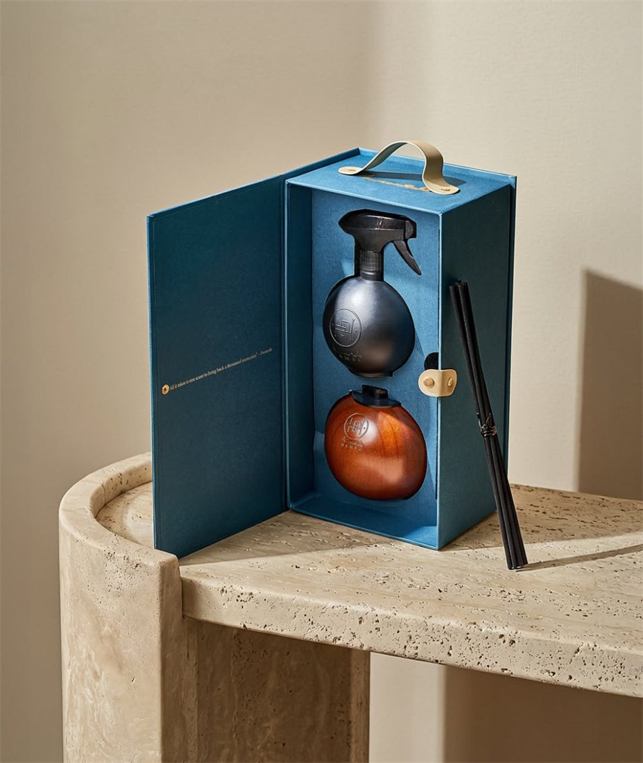 An open book-style gift box holding a black home spray and a wooden diffuser sphere with reed sticks, resting on a travertine table