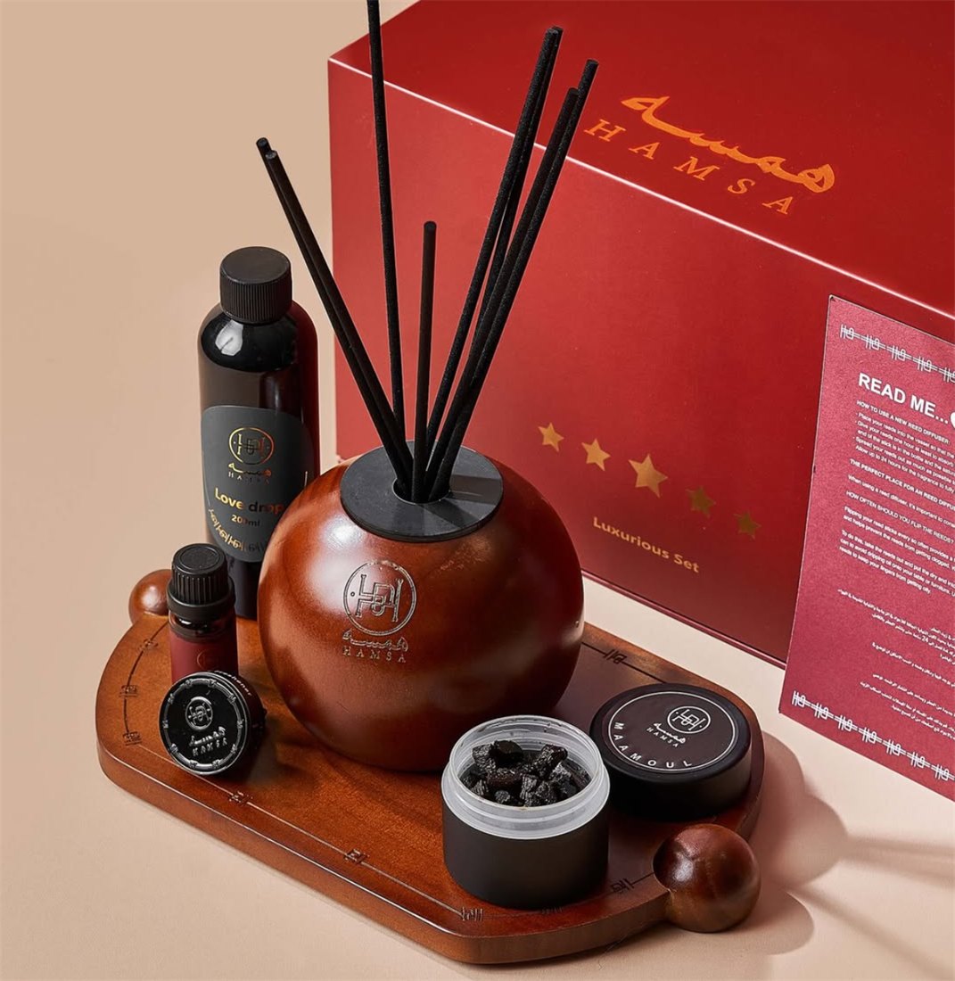 A wooden diffuser sphere with reed sticks, an oil dropper bottle, and a bakhoor jar arranged on a wooden tray in front of a red Luxurious Set gift box