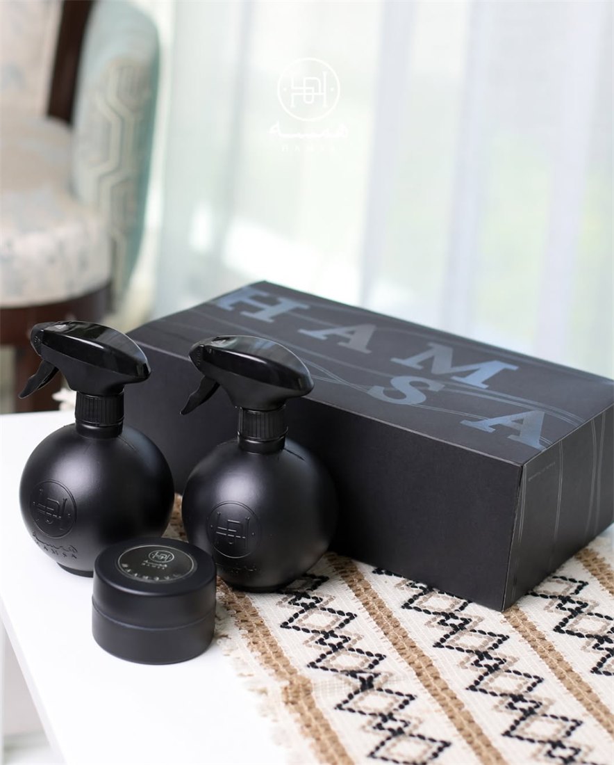 Two matte black home spray bottles and a small jar in front of a black Hamsa box on a woven table runner